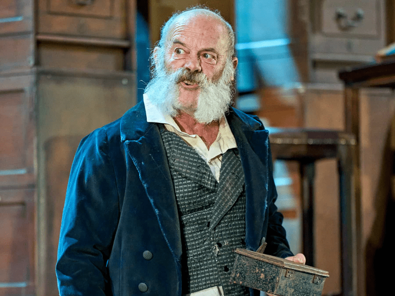 Keith Allen as Scrooge in Mark Gatiss’ A Christmas Carol. A man with bushy grey mutton chops facial hair, wearing a blue velvet jacket, holds a small cash tin and sneers.