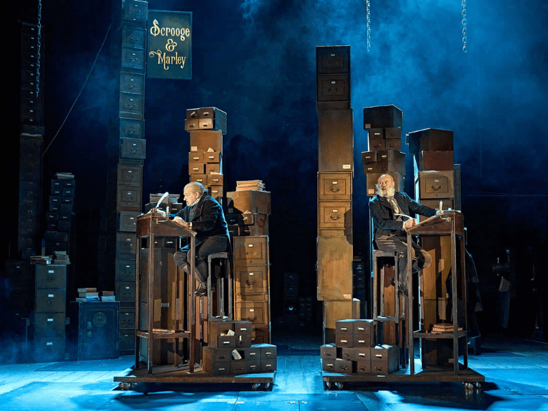 Peter Forbes as Marley and Keith Allen as Scrooge in Mark Gatiss’ A Christmas Carol. Two men sit at very tall desks holding quills, surrounded by tall filing cabinets, with a sign that says Scrooge & Marley. 