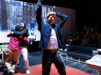 Large screen in background displaying a street scene. Two males happily embrace in background whilst one single male, wearing a demin jacket and jeans, raises a fist in the air in celebration with their other hand on their head.