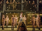 Image: Anthony Roth Costanzo stood amidst a group of people, they are all wearing golden robes and the stage is glowing golden.