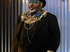 Image: Keele Watson stood on stage, wearing a black outfit with gold jewels and different gem stones and green eye makeup.