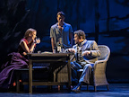 Man and woman sat on wicker chairs having a drink being overlooked by another man stood in the shadows.