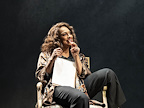 Woman sat on chair with a sketchpad in her lap and pencil in her mouth.