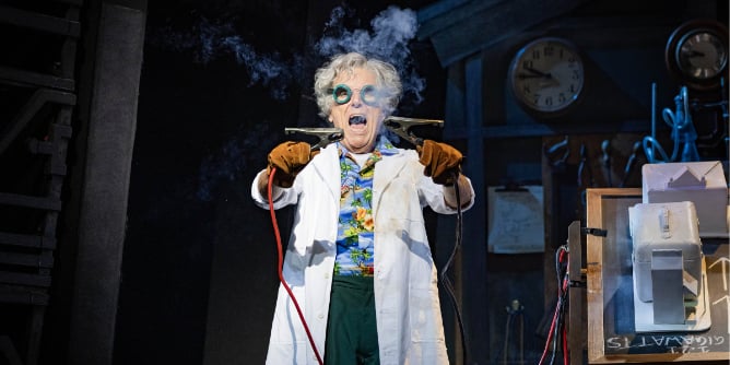 Back To The Future The Musical in London. Brian Conley stars as Doc, wearing a white lab coat and holding two wires together, which are producing smoke in front of his face.