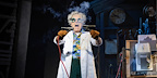 Back To The Future The Musical in London. Brian Conley stars as Doc, wearing a white lab coat and holding two wires together, which are producing smoke in front of his face.