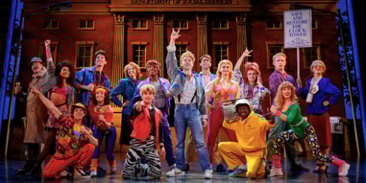 Back To The Future The Musical in London. The cast are gathered in a group in front of the town hall wearing 80s costumes, with hands in the air.