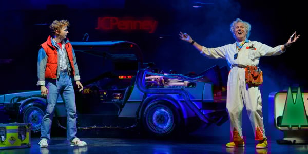 Back To The Future The Musical in London. Marty McFly, wearing jeans and a red gilet, and Doc Brown, wearing a white jumpsuit, stand across from each other with the DeLorean in between them.