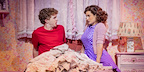 Back To The Future The Musical in London. Marty McFly, wearing a red t-shirt, and Lorraine Baines, wearing a blue dress, sit in a single bed - Marty is looking confused.