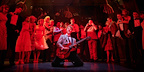 Back To The Future The Musical in London. Marty, wearing a suit, is playing the guitar the guitar on his knees, with the ensemble stood around gim wearing suits and dresses.