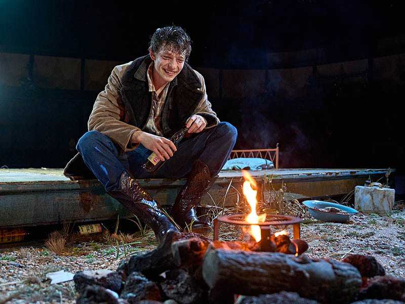 Cowboy sat in front of a fire holding a bottle of alcohol.