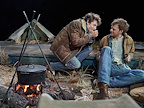 Two men sat around a camp fire. One is playing the harmonica whilst the other is watching.