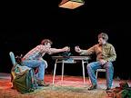 Two men sat at a table toasting each other with a drink. Back packs and cowboy hats are sat on the floor near their feet.