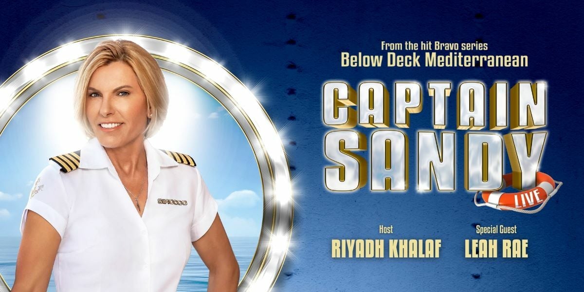 Text: From the hit Bravo series Below Deck Mediterranean, Captain Sandy Live. Image: Captain Sandy infront of a glittering porthole, the background is a sea-blue and Sandy is smiling into the camera.