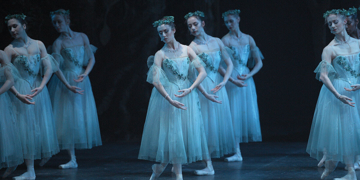 The English National Ballet. Giselle. A Woman in white poses gracefully, arms crossed, hands pointing up toward her shoulders. She is standing in a bower of flowers.
