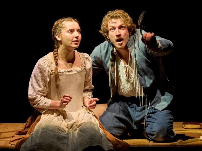 Frankie Hastings (Eliza) wearing a brown skirt, a pale green corset and a floral shirt sits next to Tow Varey (William), who is wearing blue breeches, a blue jacket and a white shirt. Tom gestures with left arm.  Photo by Manuel Harlan © RSC