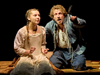 Frankie Hastings (Eliza) wearing a brown skirt, a pale green corset and a floral shirt sits next to Tow Varey (William), who is wearing blue breeches, a blue jacket and a white shirt. Tom gestures with left arm.  Photo by Manuel Harlan © RSC