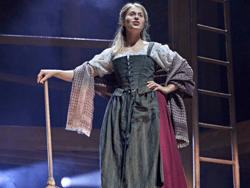 Mhairi Gayer (Shakespeare’s Landlord) stands on a wooden stage, holding a broom. Mhairi wears a maroon skirt, black corset and apron, maroon patterned shawl and cream patterned blouse. Photo by Manuel Harlan © RSC