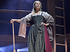 Mhairi Gayer (Shakespeare’s Landlord) stands on a wooden stage, holding a broom. Mhairi wears a maroon skirt, black corset and apron, maroon patterned shawl and cream patterned blouse. Photo by Manuel Harlan © RSC