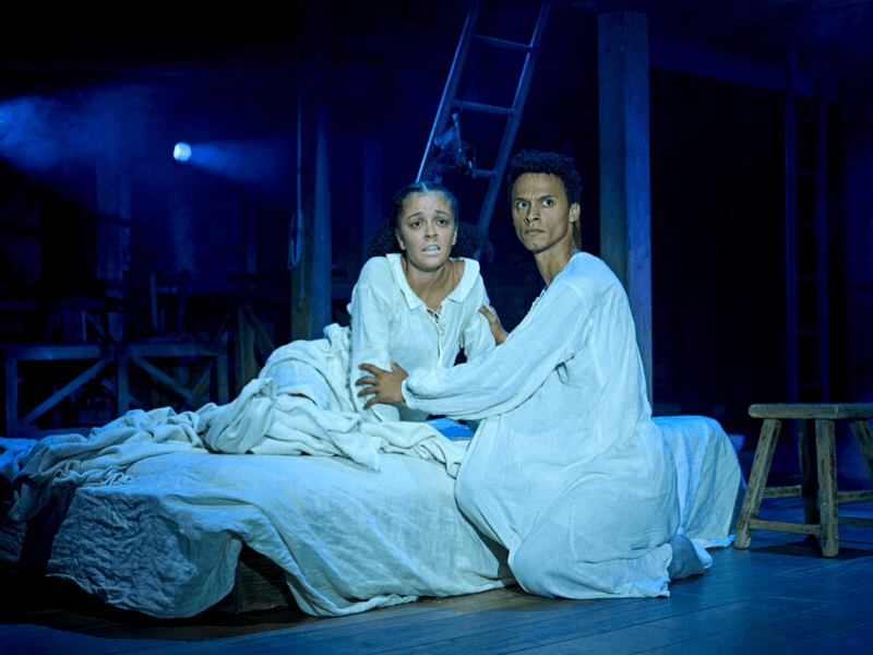 Alex Jarrett (Judith) lies on a cream bed, wearing a cream nighshirt with a face in distress. Ajani Cabey (Hamnet), wearing a cream nightshirt, looks to the left in distress.  Photo by Manuel Harlan © RSC