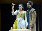 Madeleine Mantock (Agnes), wearing a yellow corset, cream blouse and yellow skirt with cream apron, holds a straw poppet. Behind Madeleine stands Gabriel Akuwudike (Bartholemew), who is dressed in a greet coat, black boots, black breeches and a black jacket.  Photo by Manuel Harlan © RSC
