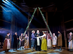 The cast of Hamnet stand on a wooden stage. Behind them, two wooden ladders fashioned into an ‘A’, draped with flowers.  Photo by Manuel Harlan © RSC