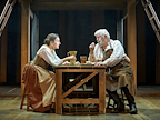 Liza Sadovy (Mary), wearing a white shirt, green corset and skirt in shades of brown, sits at a wooden table. Opposite her sits Peter Wight (John) with white hair and white beard, wearing a black waistcoat and brown apron.  Photo by Manuel Harlan © RSC