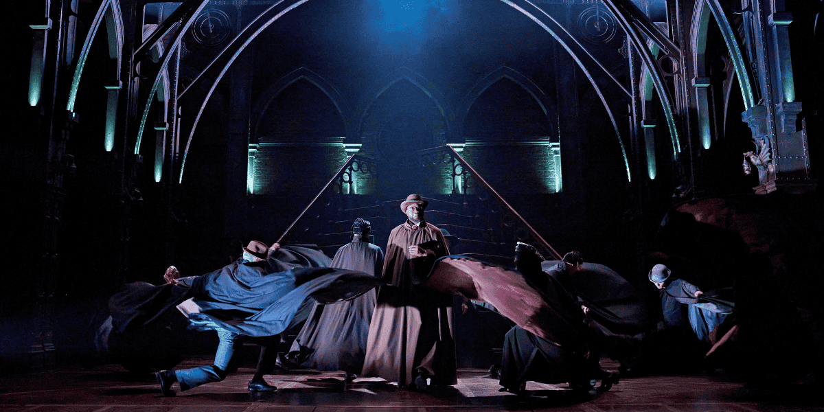 A man in brown robes and a bowler hat is stood centrestage, while others move around him dramatically swirling their robes.
