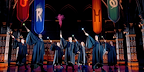 The ensemble, wearing school robes, stand in front of the school banners. They're all smiling and holding their wands up, most of which are lit up at the tip, while the boy in the middle sends up red smoke.