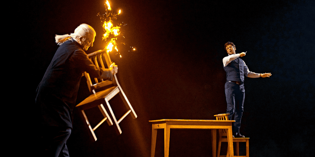 Image: Two wizards as one casts a fire spell with a wand and the other raises a chair to defend himself Show: Harry Potter and the Cursed Child