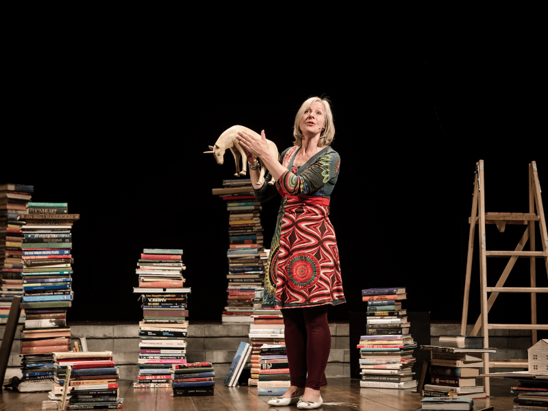 Danyah Miller on a stage surrounded by piles of books and a step ladder. holding a wooden unicorn figure