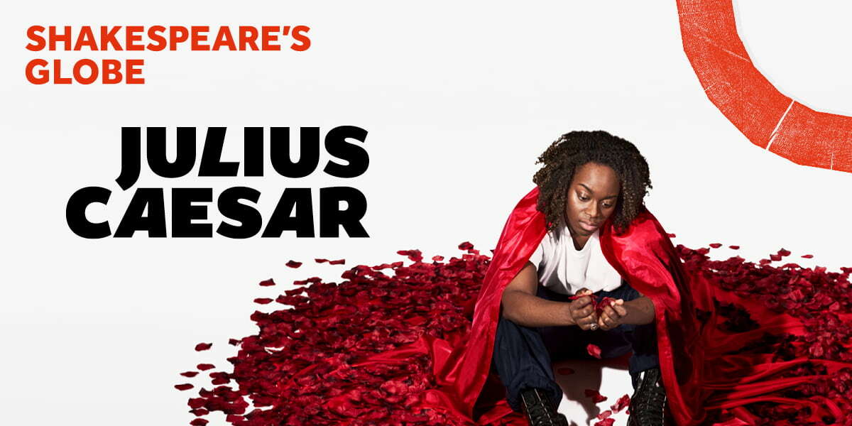 TEXT: Shakespeare's Globe Julius Caesar. An actor wearing baggy blue trousers tucked into black lace-up boots with a white shirt and red cape sits in the middle of a pile of red rose petals. The actor is holding petals in their cupped hands and looking at them sadly.
