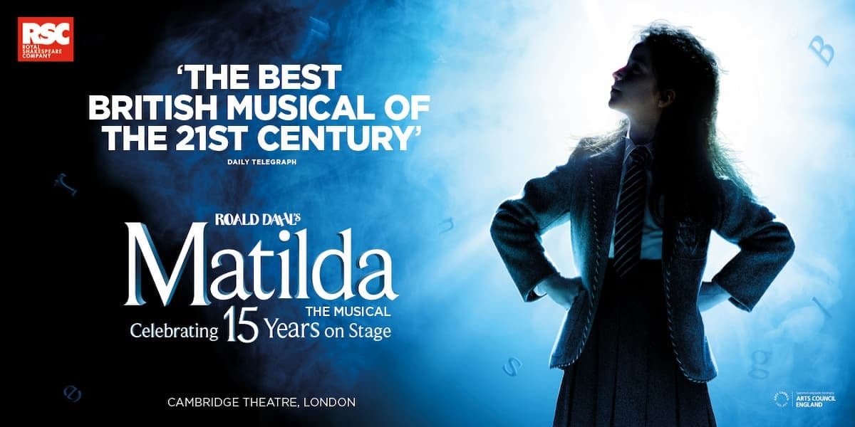 Roald Dahls' Matilda The Musical, Cambridge Theatre London. RSC Royal Shakespeare Company. Image: A little girl (Matilda) stood with her hands on her hips looking up into the left-hand distance. She is stood in-front of a glowing blue lights, which makes her look almost like a silhouette. It is a powerful image.
