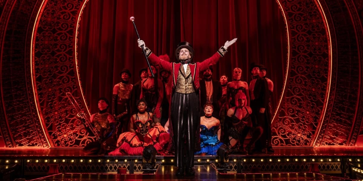 Harold Zidler in a red tailcoat stands centre stage, arms raised, with the cast behind him in Moulin Rouge! The Musical London at the Piccadilly Theatre.