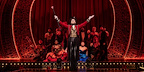 Harold Zidler in a red tailcoat stands centre stage, arms raised, with the cast behind him in Moulin Rouge! The Musical London at the Piccadilly Theatre.