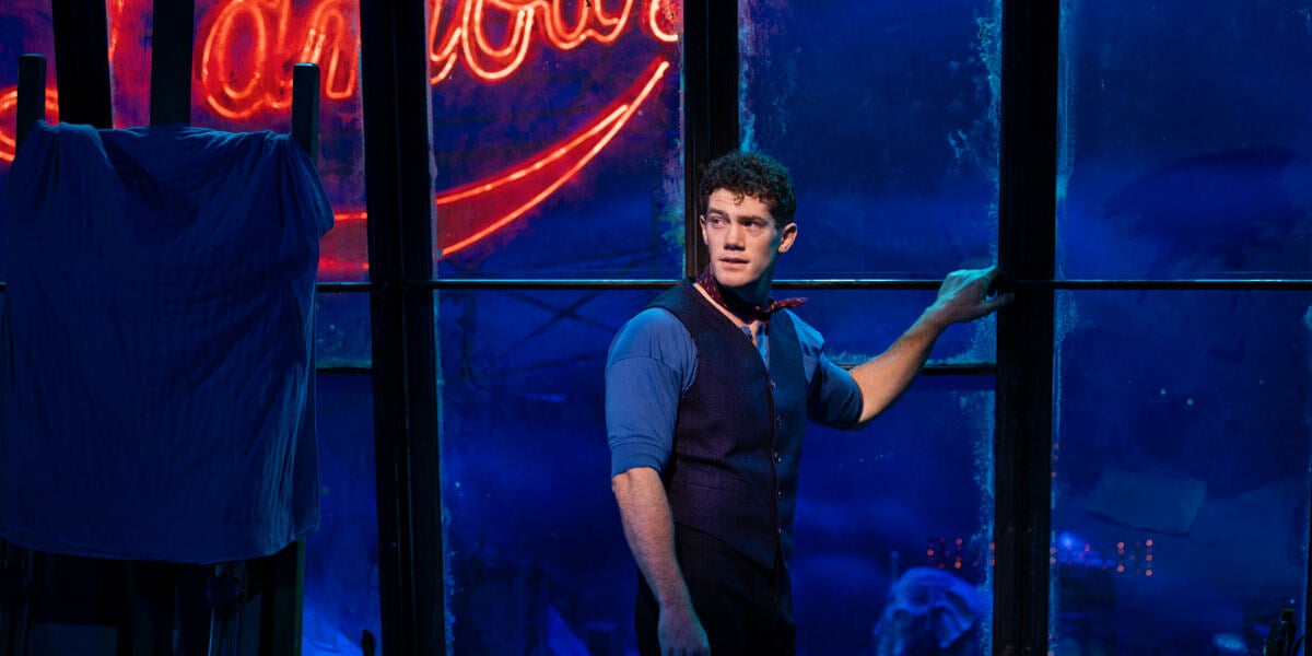 Christian, played by Alastair Brammer, stands by a blue-lit window with the Moulin Rouge sign glowing behind him in Moulin Rouge! The Musical London.