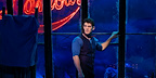 Christian, played by Alastair Brammer, stands by a blue-lit window with the Moulin Rouge sign glowing behind him in Moulin Rouge! The Musical London.