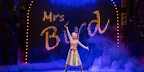 Woman standing with arms outstretched at the front of the stage with Mrs. Bird in jazzy lights lit up behind her in Paddington the Musical.