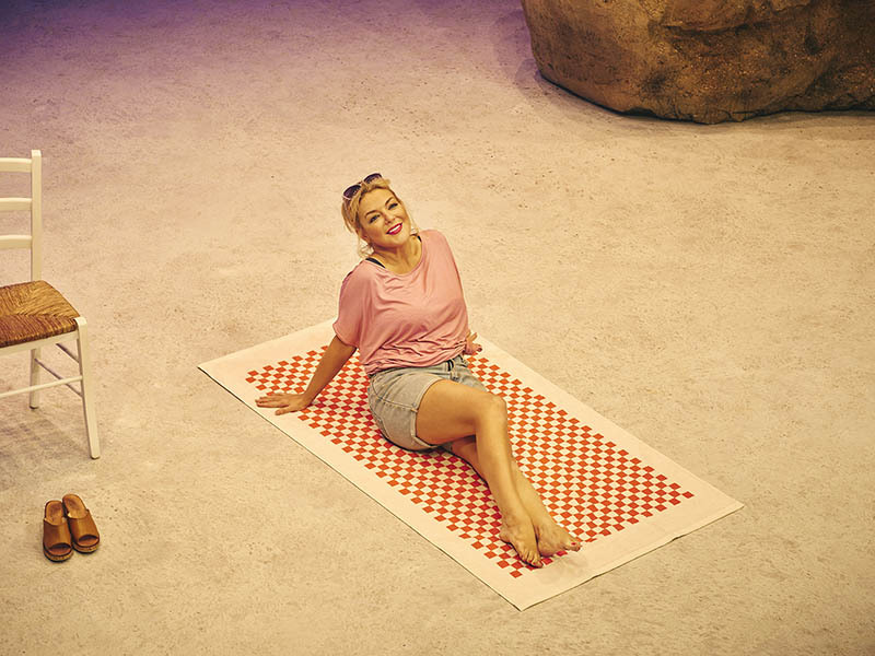 Sheridan Smith in Shirley Valentine - reclining on beach towel on beach, sunglasses on head, wearing tshirt and denim shorts.