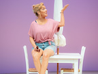 Sheridan Smith in Shirley Valentine - sat on a white table with a parasol in it wearing denim shorts and pale pink t-shirt holding a pair of sunglasses.