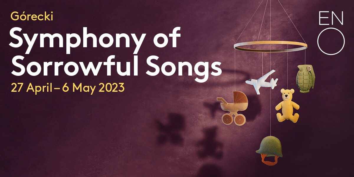Gorecki Symphony of Sorrowful Songs 27 April - 6 May 2023 ENO. A mottled purple background. A child's mobile cast shadows on the wall. Hanging from the mobile are a buggy, an aeroplane, a teddy bear, a grenade and an army helmet.