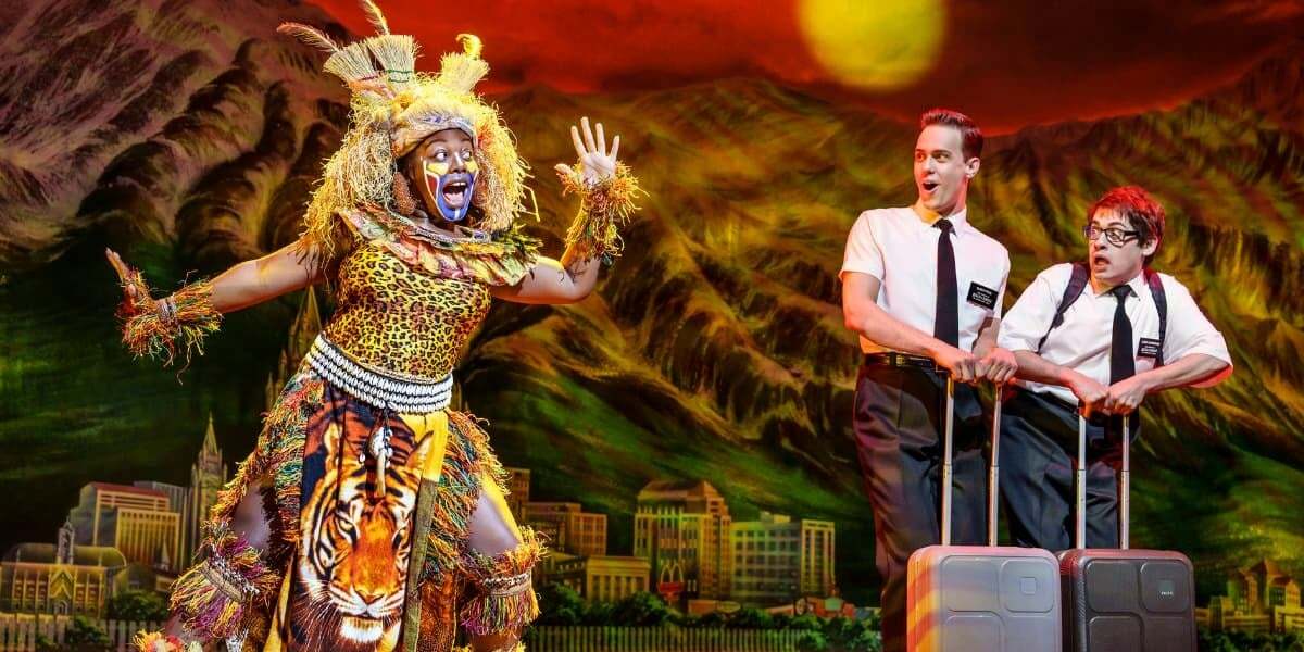 A costumed character in tribal attire gestures dramatically while two Mormon missionaries stand with suitcases in the Book of Mormon musical.