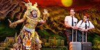 A costumed character in tribal attire gestures dramatically while two Mormon missionaries stand with suitcases in the Book of Mormon musical.