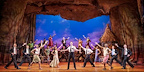 Full cast of the Book of Mormon musical perform a high-energy group number against a village backdrop.