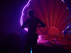 Man with his back to us stood infront of a bed with a giant clam shell headboard.