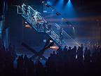 Crowd stood around a staircase bathed in blue light whilst multiple people are on the staircase in various poses.