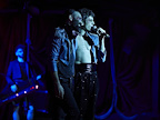 Two men, one  with an open shirt and holding a microphone.