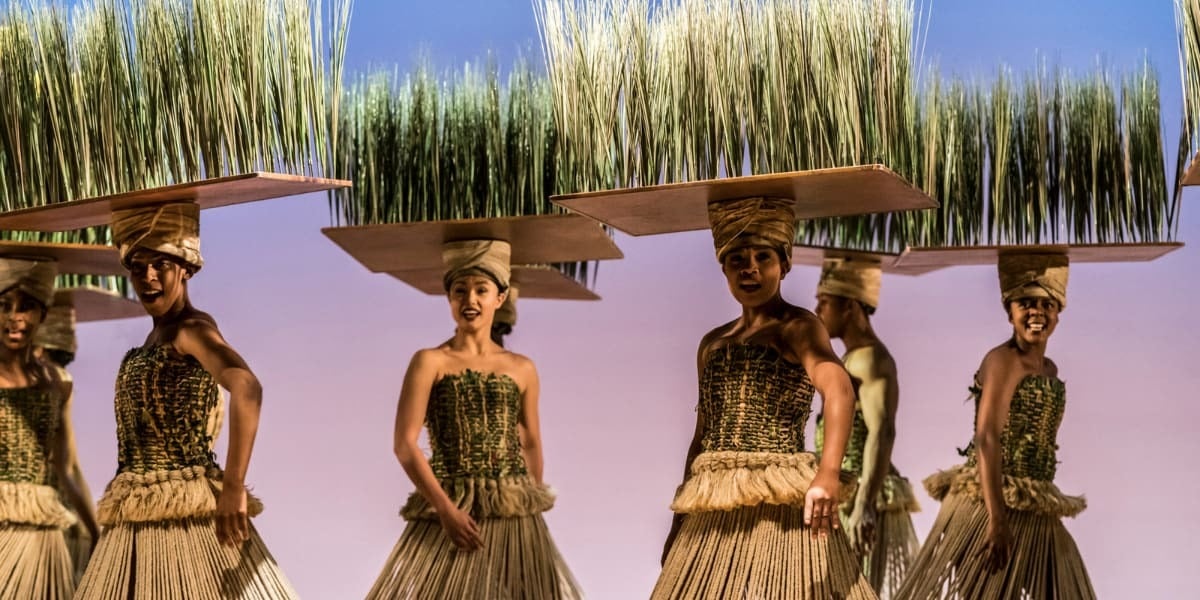 Performers carry tall grass props across the stage during the savannah scenes of The Lion King musical.