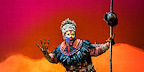 Rafiki appears on stage holding her staff, wearing vibrant costume and face paint in The Lion King musical.