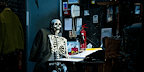 Skeleton sitting on a desk with earphones on in The Play That Goes Wrong