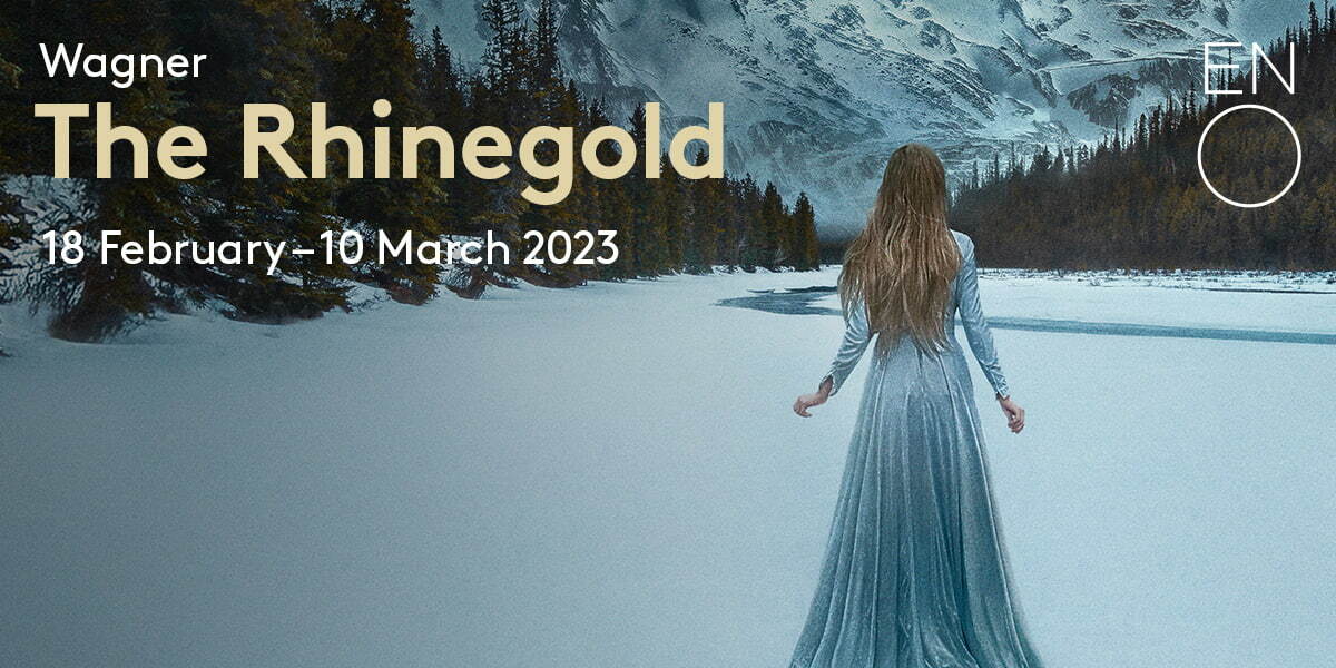 Wagner The Rhinegold 18 February - 10 March 2023 ENO. There is a snow-covered mountain in the distance. In the middle of a snowy valley flanked by pine trees a river winds. In the foreground a woman in a long-sleeved light blue dress and long blonde hair stands so we can only see her back.